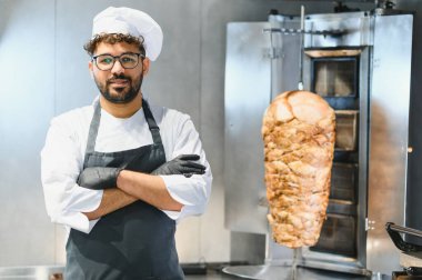 Çapraz kollarla poz veren, siyah eldiven ve önlük giyen, profesyonel bir mutfakta arka planda dönen bir shawarma tükürüğü olan kendine güvenen bir şef.