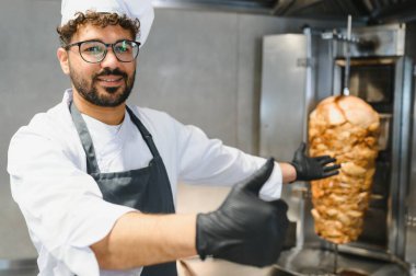 Profesyonel aşçı siyah eldiven giyer. Lokanta mutfağında kızarmış shawarma eti sunarken baş parmak hareketi yapar.