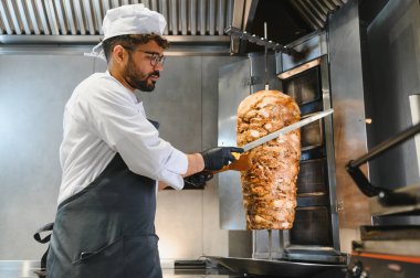 Profesyonel bir şef, ticari bir mutfakta lezzetli shawarma etini dönüşümlü bir tükürükten ustalıkla dilimliyor. Popüler bir sokak yemeği hazırlıyor.
