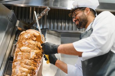Profesyonel bir şef, büyük bir bıçak ve spatula kullanarak, ticari bir mutfakta dönen bir tükürükten lezzetli shawarma etini ustalıkla dilimliyor.