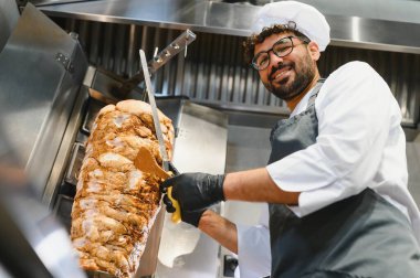 Siyah eldiven ve önlük takan profesyonel bir şef, ticari bir mutfakta şiş şiş eti ustalıkla dilimliyor, popüler bir sokak yemeği hazırlıyor.