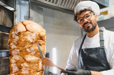 Profesyonel bir mutfakta, üniforma ve eldiven giyerek shawarma ya da donör kebabı için dönen bir tükürükten et dilimleyen Ortadoğulu şef.
