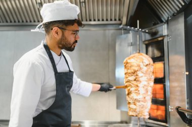 Profesyonel bir aşçı, eldiven ve önlük giyerek, ticari bir mutfakta dönen bir shawarma tükürüğünden et doğruyor.