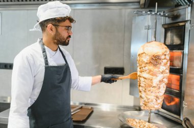 Profesyonel bir aşçı, ticari bir mutfakta çalışıyor. Dönen bir shawarma tükürüğünden et kesmek için ahşap bir spatula kullanıyor.