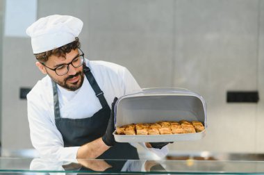Profesyonel aşçı, siyah eldiven ve üniforma giyiyor. Elinde bir tepsi taze pişmiş baklava var. Doğu restoranının mutfağında.