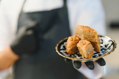 Şefin siyah eldiven giymesi zarif bir tabakta baklava porsiyonları sunması Orta Doğu ya da Akdeniz mutfağında uzmanlığı vurguluyor.