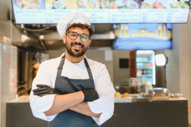 Kollarını kavuşturmuş, siyah eldivenler ve önlükle modern bir ticari mutfakta dijital menüde poz veren kendine güvenen bir şef.