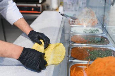 Siyah eldiven giyen aşçı bir fast food restoranında pide ve çeşitli malzemelerle shawarma ya da kebap dürümü hazırlıyor.