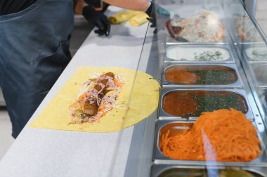 Aşçı, bir fast food restoranının arka planında soslar ve sebzelerle birlikte shawarma ambalajına malzeme ekliyor.