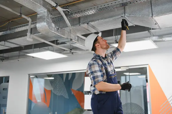 Güvenlik kaskı takan bir inşaatçı modern bir ofiste tamirat sırasında havalandırma borularını kontrol ediyor.