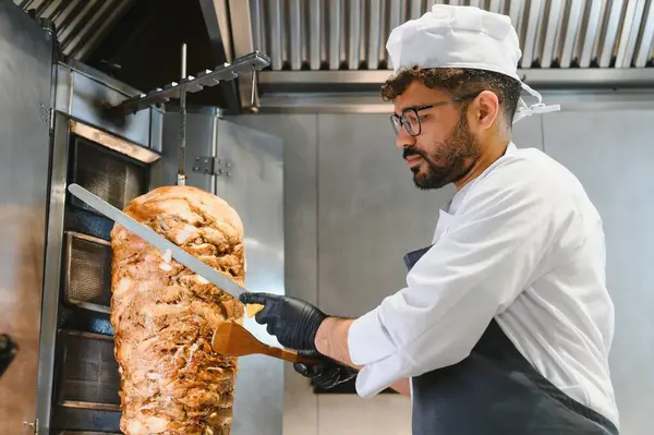 Profesyonel bir şef, ticari bir mutfakta lezzetli shawarma etini dönüşümlü bir tükürükten ustalıkla dilimliyor. Popüler bir orta doğu sokak yemeği hazırlıyor.