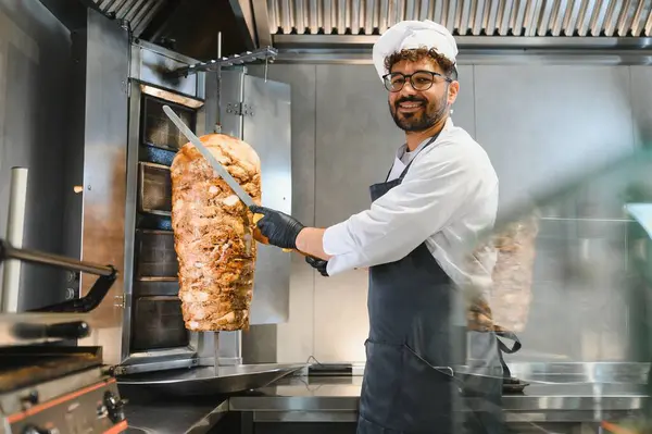 Profesyonel bir şef, ticari bir mutfakta lezzetli shawarma etini dönüşümlü bir tükürükten ustalıkla dilimliyor. Popüler bir sokak yemeği hazırlıyor.