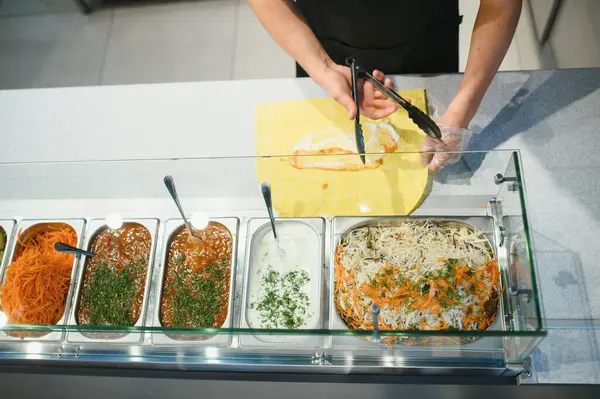 Aşçı pide ekmeğe et koymak için maşa kullanıyor, ön planda soslar ve sebzeler var.