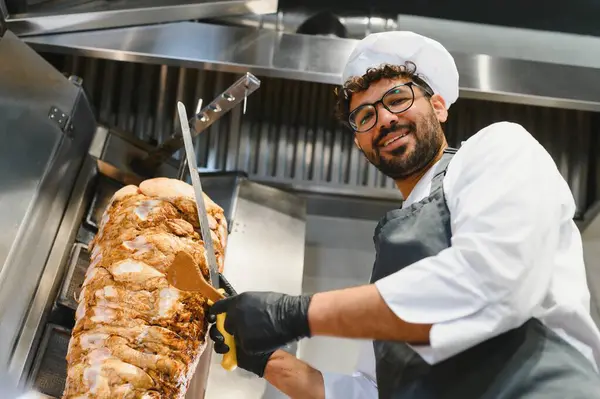 Gülümseyen şef profesyonel mutfakta büyük bir bıçakla shawarma eti dilimliyor, geleneksel orta doğu yemeğini hazırlıyor.