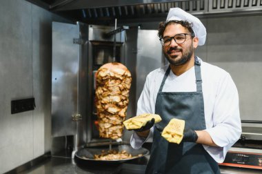 Aşçı siyah eldiven ve önlük takıyor. Restoran mutfağında iki parça shawarma tutuyor. Dönüşümlü donör kebabının yanında.