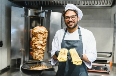 Gülümseyen şef, yeni yapılmış iki shawarma 'yı sunar. Arka planda da bir döner döner kebap var.
