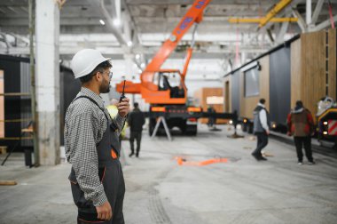Arap mühendis prefabrik ev üretim hattında telsiz koordine operasyonları yapıyor, vinç ve işçiler arka planda.