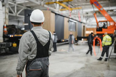 Büyük endüstriyel fabrikada prefabrik modüler ev üretimini denetleyen kasklı ve tulumlu inşaat işçisi.