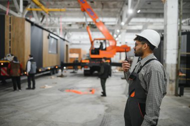 Arap mühendis güvenlik kaskı takıyor, telsiz kullanıyor ve büyük inşaat atölyesinde prefabrik ev yapımını denetliyor.