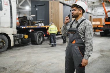 Modüler ev yapım atölyesinde telsiz kullanarak güvenlik kaskı takan Arap inşaat işçisi operasyonları koordine ediyor ve sorunsuz bir iş akışı sağlıyor.