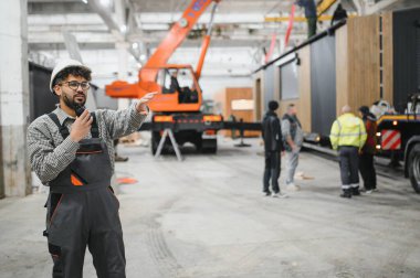 Üniforma giyen Arap mühendis telsizle iletişim kuruyor ve modüler ev yapımı atölyesinde vinç operasyonlarını koordine ediyor.