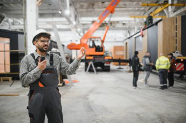 Üniforma giyen ve telsiz kullanarak iletişim kuran Arap mühendis büyük bir inşaat atölyesinde modüler ev toplantısını koordine ediyor.