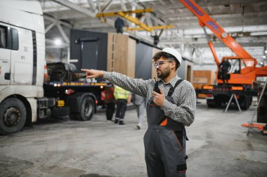 Güvenlik kaskı takan ve telsizi elinde tutan Arap mühendis modüler bir ev üretim tesisini işaret ediyor.