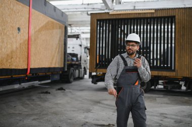 Güvenli kask takan Arap inşaat işçisi, büyük bir inşaat atölyesinde modüler ev yapımı sırasında telsiz kullanarak iletişim kuruyor.