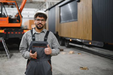 Güvenlik kaskı takan ve elinde telsiz olan Arap inşaat işçisi modüler ev yapımı atölyesinde bir jest yapıyor.