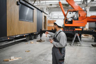 Arap mühendis telsiz kullanıyor ve büyük inşaat atölyesinde prefabrik modüler ev üretimini denetliyor.