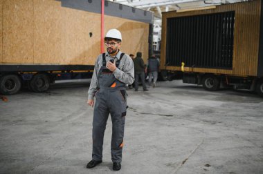 Güvenlik kaskı ve tulum giyen Arap mühendis telsiz kullanıyor, bir inşaat atölyesinde modüler ev ulaşımını koordine ediyor.