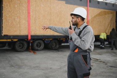İnşaat işçisi güvenlik kaskı takıyor ve modüler ev teslimatını düz kamyon ile yönetirken telsiz kullanıyor.