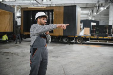 Büyük bir atölyede prefabrik modüler ev üretimini denetlerken güvenlik kaskı takan ve üniforma giyen Arap inşaat işçisi