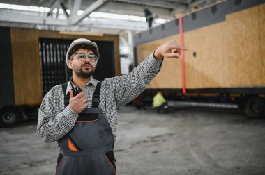 İnşaat işçisi telsiz kullanıyor ve fabrikadaki modüler ev ulaşımını denetlerken işaret ediyor.