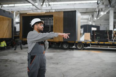 Güvenlik kaskı takan Arap mühendis ve iş tulumları büyük bir inşaat atölyesinde modüler ev üretimini gösteriyor ve denetliyor.