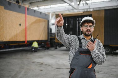 Arap mühendis telsiz kullanıyor ve büyük inşaat atölyesinde modüler ev üretimini denetlerken işaret ediyor.