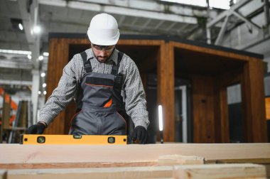 Odaklanmış inşaat işçisi ahşap kiriş seviyesini ruh seviyesiyle kontrol ediyor, büyük marangozluk atölyesinde modüler ev yapımı üzerinde çalışıyor.