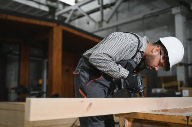 Yoğunlaşmış marangoz, ahşap bir kalas üzerinde kablosuz matkap kullanıyor, bir inşaat atölyesinde prefabrik ev yapısı üzerinde çalışıyor.