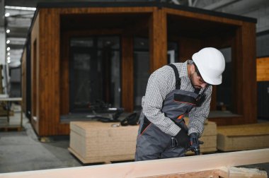 Yetenekli marangoz, modern atölyede prefabrik modüler ev bölümü inşa etmek için ahşap kalas üzerinde elektrikli matkap kullanır.