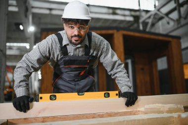 Arap inşaat işçisi, prefabrik ev atölyesinde ahşap kalas üzerinde doğru ölçümler yapmak için ruh seviyesini kullanıyor.