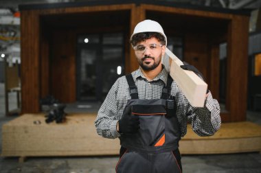 Kendine güvenen inşaat işçisi, ahşap kalas taşıyor, prefabrik modüler konutlarda uzmanlık gösteriyor.