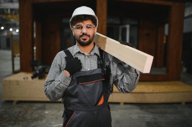 Arap inşaat işçisi omuzunda ahşap kalas taşıyor, modern atölyede prefabrik ev üzerinde çalışıyor.