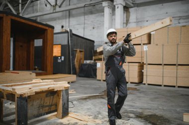 Marangoz güvenlik kaskı ve eldiven takıyor. Modüler bir inşaat atölyesinde ahşap tahta taşıyor.