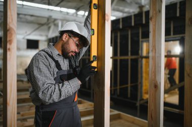 Prefabrikasyon atölyesinde modüler ev montajı sırasında ahşap kirişin tam olarak dikey hizalanmasını sağlamak için ruh seviyesini kullanan odaklanmış inşaat işçisi