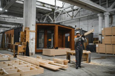 Marangoz ahşap kalas taşıyor. İnşaat malzemeleri ve modüler ev yapılarıyla çevrili prefabrik ev atölyesinde çalışıyor.