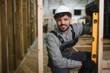 Gülümseyen Arap inşaat işçisi ahşap kiriş üzerinde ruh seviyesini kullanarak, bir atölyede modüler ev yapımı sırasında dikey hizalamayı garanti ediyor