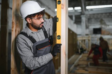 Odaklanmış inşaat işçisi ruh seviyesini kullanarak ahşap kirişin dikeyliğini kontrol ediyor, marangozluk atölyesinde prefabrik ev yapısı üzerinde çalışıyor.