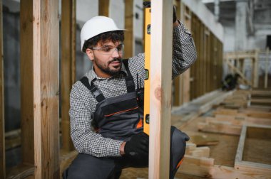 Arap inşaat işçisi, bir atölyede modüler ev inşaatı sırasında ahşap kirişin dikey hizalanmasını sağlayan bir ruh seviyesi kullanıyor.