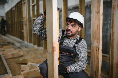 Arap inşaat işçisi, bir modüler ev üretim atölyesi içindeki ahşap kirişin tam olarak dikey hizalanmasını sağlayan bir ruh seviyesi kullanıyor.