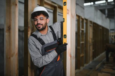 Arap inşaat işçisi prefabrik ev atölyesindeki ruh seviyesini kullanarak ahşap kirişin dikeyliğini kontrol ediyor.